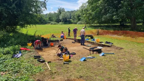 A team of archaeologists works in a hole in a grassy field. It is sunny. 