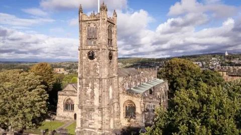 Historic England A church with a tower and clock seen on two sides has mullioned windows on the either side of the building. The church is surrounded by trees. Some houses and hills can be seen in the distance.