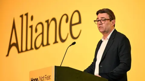 PA Media Andy Burnham stands up on stage in front of a large yellow Alliance banner. He is stood at a podium with a singular long microphone. He has short, brown hair and is wearing a dark jacket and a white shirt.