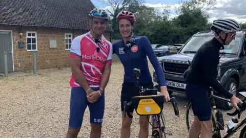 Jill and Phil are both wearing cycling gear and helmets and stand beside one another in a gravel-covered area with a building in the background. Jill is holding her bike and a third person in cycling gear is standing on the right hand side of the image.
