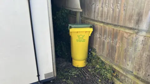 Luke Deal/BBC A yellow bin with a green lid sits in between a wooden fence and white pub. It has been chained to a post behind it. 