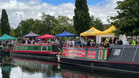 Narrowboats are moored beside a canal as visitors browse colourful stalls and displays at a canal-side festival.