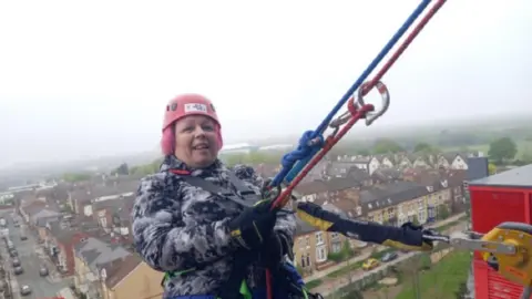 A woman wearing a pink helmet, a coat and a harness holds on to two ropes as she abseils down a building, with houses, streets and parked cars visible below her.