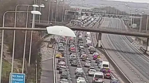 National Highways Elevated camera view of four lanes of traffic on the M25 with cars crawling or at a standstill. The opposite carriageway is entirely empty.