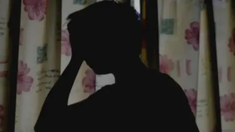 PA Media A silhouette of a person holding their head in one hand in a darkened room with the curtains drawn.