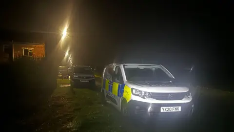 Two police vehicles are parked on a grassed verge. A red brick house is visible on the left of the image and a number of illuminated street lights are visible in the distance.