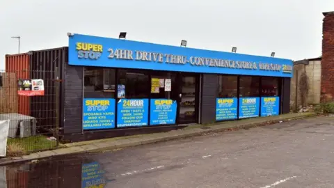 A vape and convenience shop built inside two shipping containers, with bright blue signage. The shop is in the corner of a car park, with a large puddle on the ground in front of the store. 