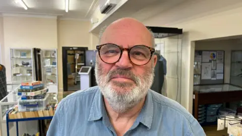 A bald man with thin grey hair on the side of his head. He has a grey beard and moustache and is wearing brown glasses. He is wearing a blue denim-type shirt. Behind him are items on display at the museum.