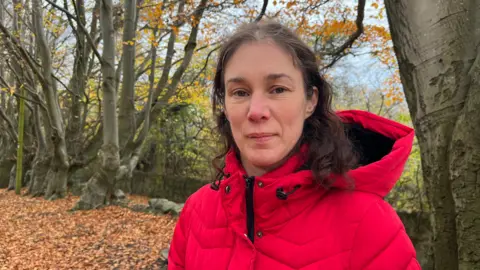 A woman in a red winter jacket with dark curly hair is looking at the camera. Behind her there are trees with yellow and orange leaves and the ground is covered with orange fallen leaved.