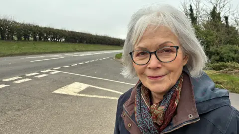 Woman with glasses smiling slightly by a road with junction markings