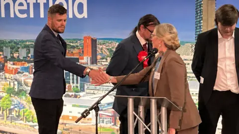A man in a blue suit shakes the hand of a woman, the counting officer, who congratulates him.