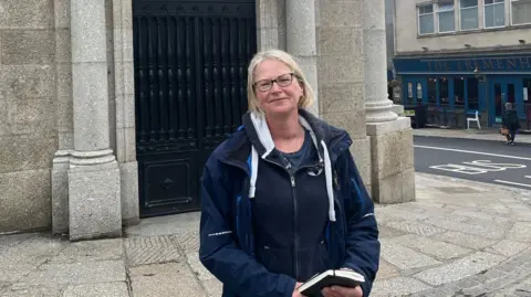 A woman with blonde hair and a blue jacket stands in front of a bank building in Penzance town centre. 