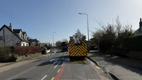 Two fire engines are parked on a road between the houses on a sunny day.