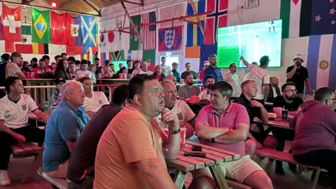Devizes Town FC A group of football fans watching a game at Devizes Town Football Club. There is a group of men sat around a bench at the front of the frame. One man is sat watching the screen while those around him are chatting. Flags from around the world are hung up around the room.