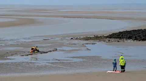 Wirral Coastguard Rescue Team A wide shot of four members of HM Coastguard Search and Rescue, dressed in yellow and blue high-visibility jackets, stand facing the muddy area of Leasowe Bay where a rescue is taking place on the beach. A large expanse of sand reaches out to the sea.