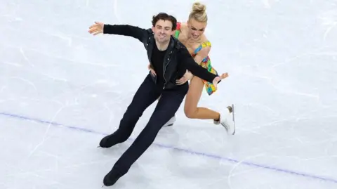 PA Media A pair of figure skaters on an ice rink. One is wearing a black leather jacket, while the other is wearing a colourful dress. Both are smiling.