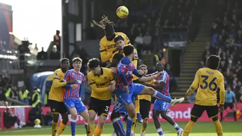 Reuters A group of footballers are mid-game on a pitch. Some players are wearing gold and black while others are wearing red and royal blue striped kit. The ball is in the air and a Wolves player is trying to header it. There are crowds blurred in the stands in the background. 