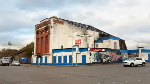 A side angle of the art deco style building. The central section has three tall windows framed by reddish-brown stonework, and all ground-level entrances are covered with metal shutters, it also shows the side profile of the metal frameword roof.