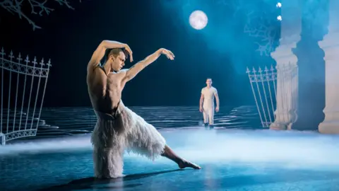 Theatre Royal Plymouth A scene from Swan Lake. A male dancer rests on one knee with his left leg outstretched and his arms curled around his head. He is bare chested and wears white feather trousers. Behind him is a man in a white T-shirt and trousers standing looking at him under a full moon. The set features wrought iron gates and part of a large stone building partly obscured by a low mist.