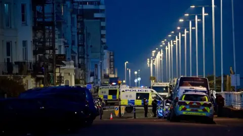 Police vans and cars are seen parked on a street with officers also standing nearby and police tape visible. There are houses on the left of the image and on the right are street lights lit up against a dark sky.