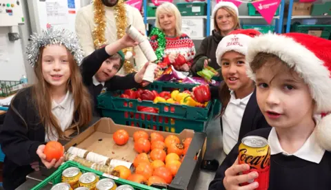 Coleham Primary School Four school pupils wearing Santa hats or tinsel holding food and standing next to boxes of Coca Cola cans, tomatoes and peppers. In the background are three adults in Christmas jumpers