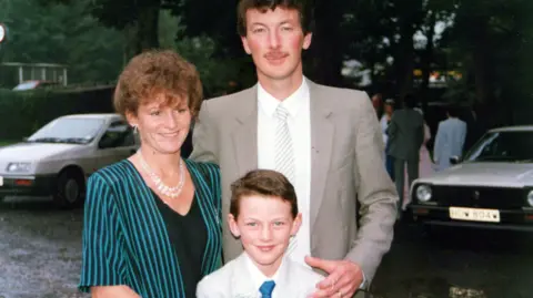 Luke Evans Luke Evans en tant que jeune garçon avec ses parents Yvonne et David. Luke porte un costume gris pâle et une cravate bleue. David porte un costume gris, une chemise blanche et une cravate rayée à droite, et Yvonne porte une veste à fines rayures bleues et noires et un haut noir, avec des perles autour du cou.