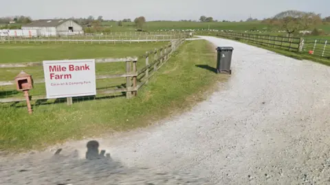 The entrance to a caravan park on farmland with a red-on-white sign reading Mile Bank Farm Caravan and Camping Park, next to a hardcore driveway edged by a wheelie bin and wooden fences, with fields of cattle in the distance.