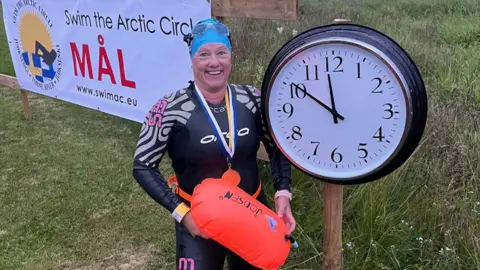 Roz Rapp standing in a wet suit holding a yellow float next to a clock with the time 11:50pm on the clock. She is smiling with medals around her neck and is in front of a sign which says "swim the arctic circle".