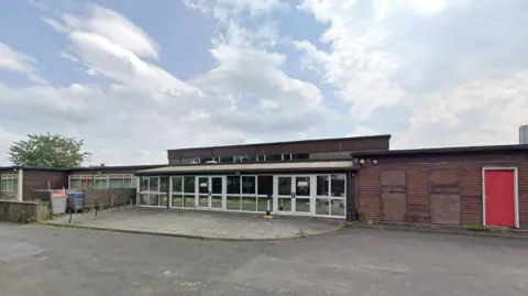 The school building is mainly single-storey, with a two storey section rising in the back. The building in the middle is lined with windows and two sets of glass doors. There is a wooden building on the right, with a red door, and a bin storage area on the left. The site looks rundown.
