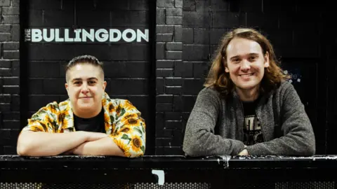 Hannah Bruce and James Taylor are leaning on a black metal bar. She is on the left wearing a flowery short and has short hair. He is on the right, has long hair and is wearing a grey jumper. Behind them is a black wall with 'The Bullingdon' in white writing.