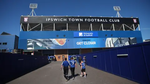 A view of Portman Road stadium from the outside, showing a group of Ipswich Town fans walking away from the camera, towards the ground. Ipswich Town Football Club is in white text across the top of the stand, which is topped by floodlights left and right.
