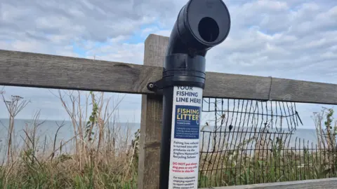 BBC / Natalie Glanvill A black fish hook bin attached to a wooden fence. There is long grass around the fence.