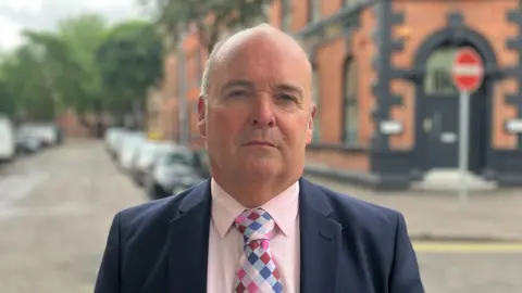 A white man looks directly into the camera unsmiling. He is balding with short grey hair on the sides of his head. He is wearing a navy formal blazer, a pink shirt and a multi-coloured checked tie. The background is blurred but he is standing on an empty street with a red brick building behind him and a number of parked cars.