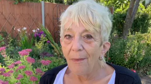 A picture of the head and shoulders of a woman with blond hair with the fence, trees and flowers of her garden behind her.