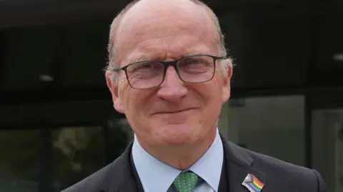 Nikki Fox/BBC Nick Hulme is wearing a grey suit. He has a green tie on and is wearing a rainbow-coloured badge that represents LGBQT plus inclusion.