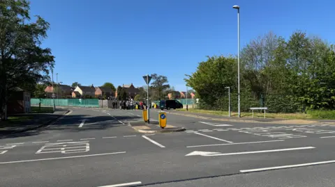 The view of the site from Packington Hill shows the junction with Pritchard Drive. Road markings instruct drivers to keep clear. A pedestrian island is in the middle of Pritchard Drive. In the distance, campaigners can be seen gathering outside the entrance to the site.