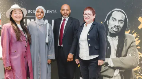 Birmingham City University Press Office A group of three women and one man stand in front of a black backdrop. They are all smartly dressed in suit jackets or long dresses. On the black backdrop there is a black-and-white photo of a man with long dreadlocks and a suit jacket.