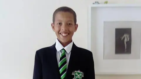 Pacemaker Noah is smiling in front of a white wall with a picture behind him. He is of mixed race and has brown short hair and brown eyes. 
He is wearing his school uniform of a black blazer with green badge, a white shirt and a green and black striped tie.