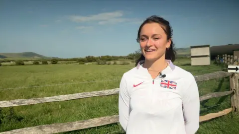 BBC Gemma Tutton, wearing a white GB athletics top, stands beside a farm gate, with a field in the background.