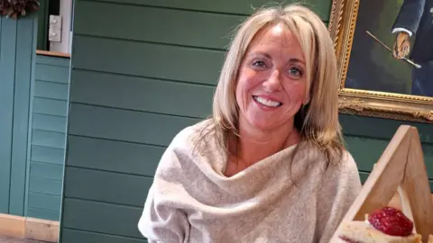 A woman with long blonde hair and wearing a grey top, smiles as she sits at a table in a restaurant.