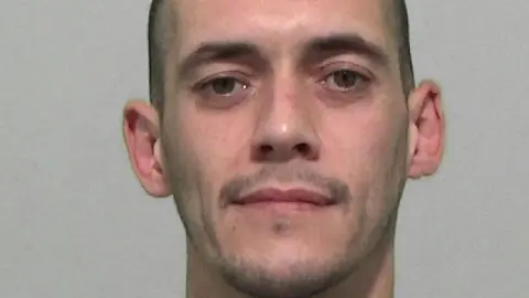 Mugshot of a man with short dark hair and stubble