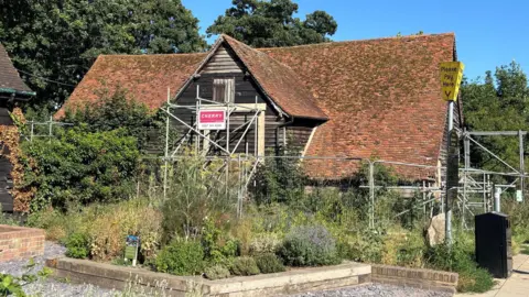 Katy Lewis/BBC A ground level view of the barn shows it surrounded by metal fences and scaffolding, and overgrown shrubbery. The roof is red with lots of moss growing on it.