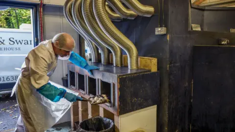 Supplied A man wearing a hairnet and white overalls and blue gloves putting what looks like salmon into a large oven