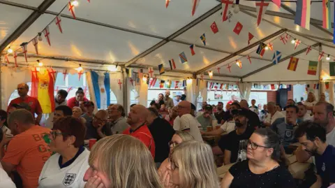 BBC / Zac Sherratt People sit inside a marquee decorated with England flags and international bunting while watching a football match on a screen out of view.
