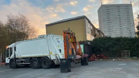 A white bin lorry is parked in an empty concrete car park. Behind it is a hedgerow, a row of houses and a block of flats.