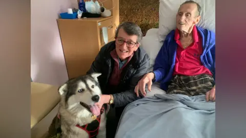 Michele Bates A dog sits with its tongue out and wearing a harness. A woman with a black jacket and glasses is holding onto the dog and leaning forward. Next to her in the hospital bed is a man wearing a blue cardigan, bright T-shirt and checked trousers.