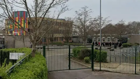 A Google Maps image of the outside of The Henry Cort Community College - the gates of the college can be seen and the college is in the distance. It has a section of the building which is multi-coloured