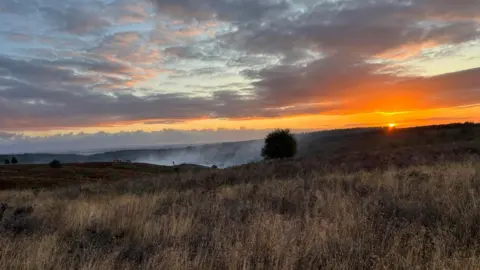 The sun is rising or setting behind the distance hillside as smoke rises from a fire in the middle of a large area of heathland.