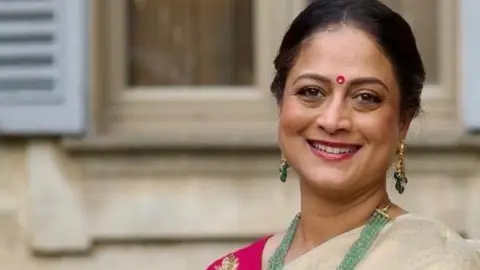 Cathy George Cathy, a woman, wearing a golden saree and pink blouse and green beaded necklace, smiling for camera.