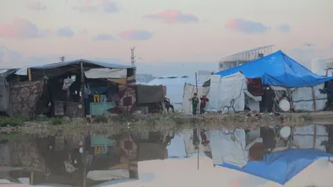 Anadolu via Getty Images Tents - one of them blue with two children outside it - are reflected in floodwater in Nuseirat refugee camp, central Gaza (17 December 2025)